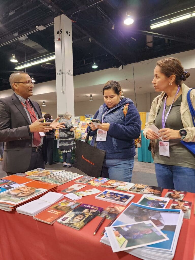 Haimdat Sawh, oficial de desarrollo de CNEWA, habla con dos trabajadores parroquiales en el estand de CNEWA en el Congreso de Educación Religiosa de Los Ángeles en Anaheim, California.