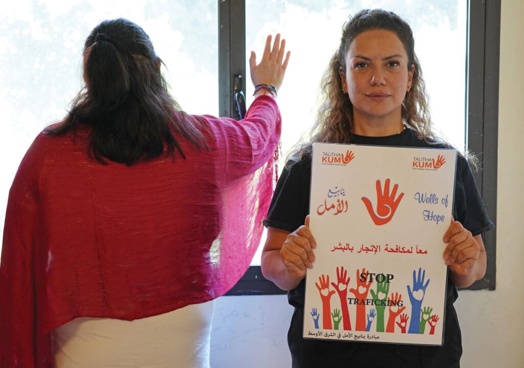Nayiri Arslanian, trabajadora social de Fuentes de Esperanza en el Líbano, sostiene un cartel contra la trata, junto a un sobreviviente de la trata que está de espalda.