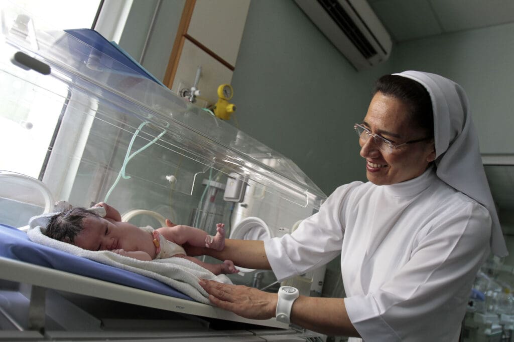 Hermana religiosa con hábito blanco sonríe a un recién nacido en una incubadora.