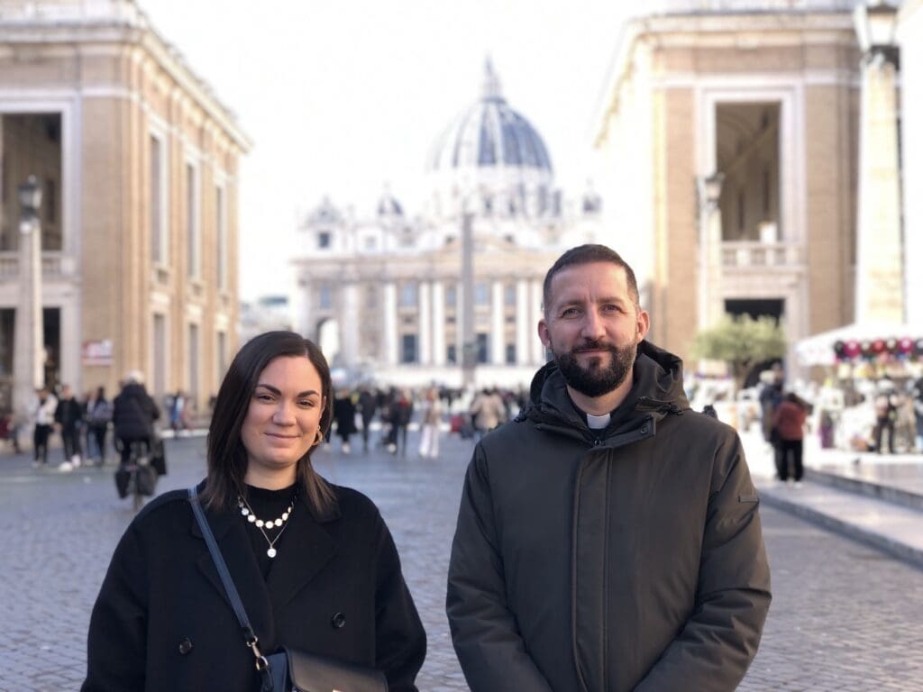 Una mujer y un sacerdote parados en la calle con la vista del Vaticano detrás de ellos.