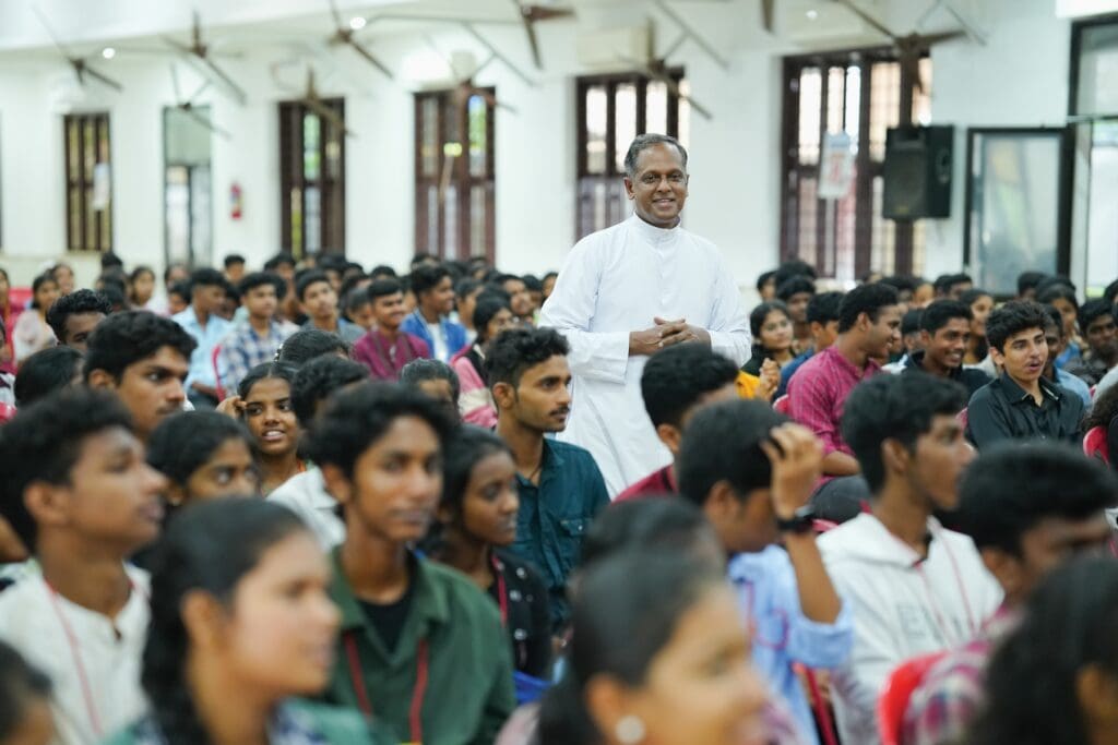 Sacerdote con sotana blanca, parado, en medio de muchos jóvenes sentados en un aula.