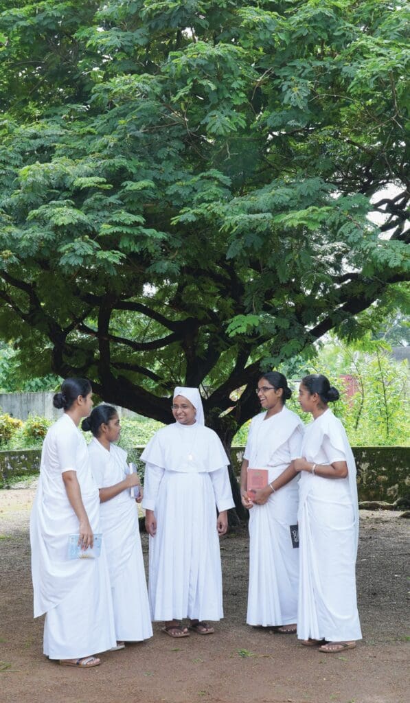 Cinco mujeres vestidas de blanco delante de un frondoso árbol. Una hermana religiosa, en el medio, y cuatro novicias.