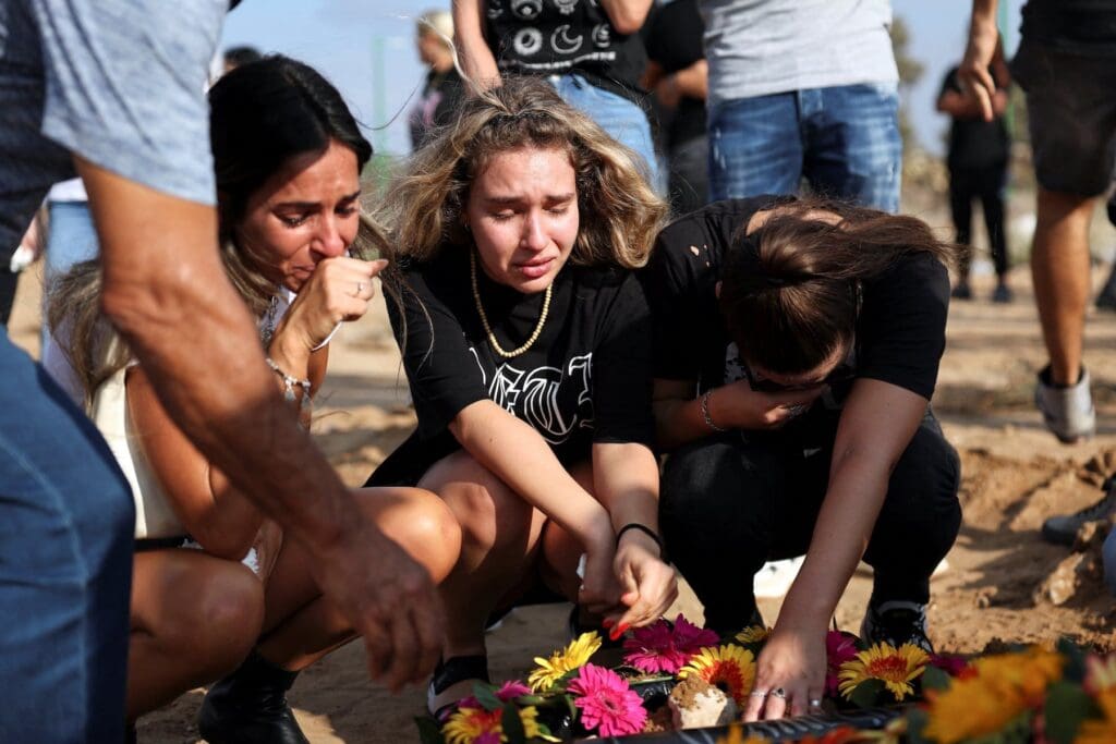tres mujeres agachadas lloran sobre una tumba cubierta de flores.