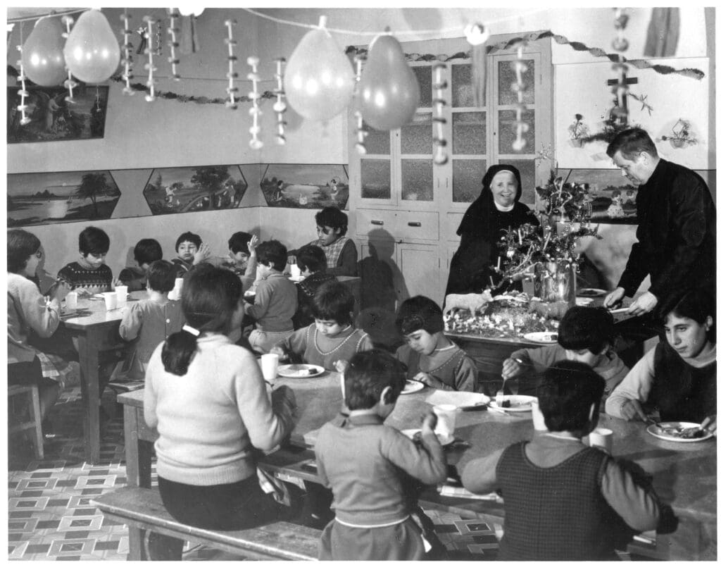 Foto de archivo en blanco y negro de un sacerdote y una hermana religiosa sirviendo en un comedor de niños.
