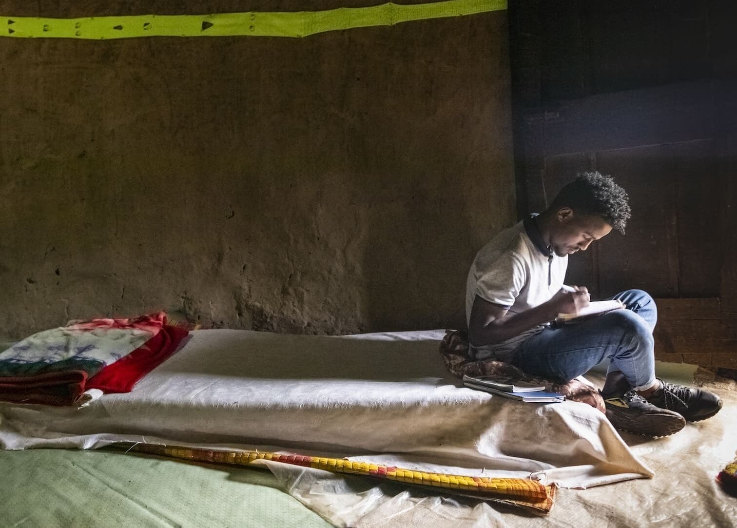 Un ragazzo seduto su un materasso poggiato a terra sta studiando.