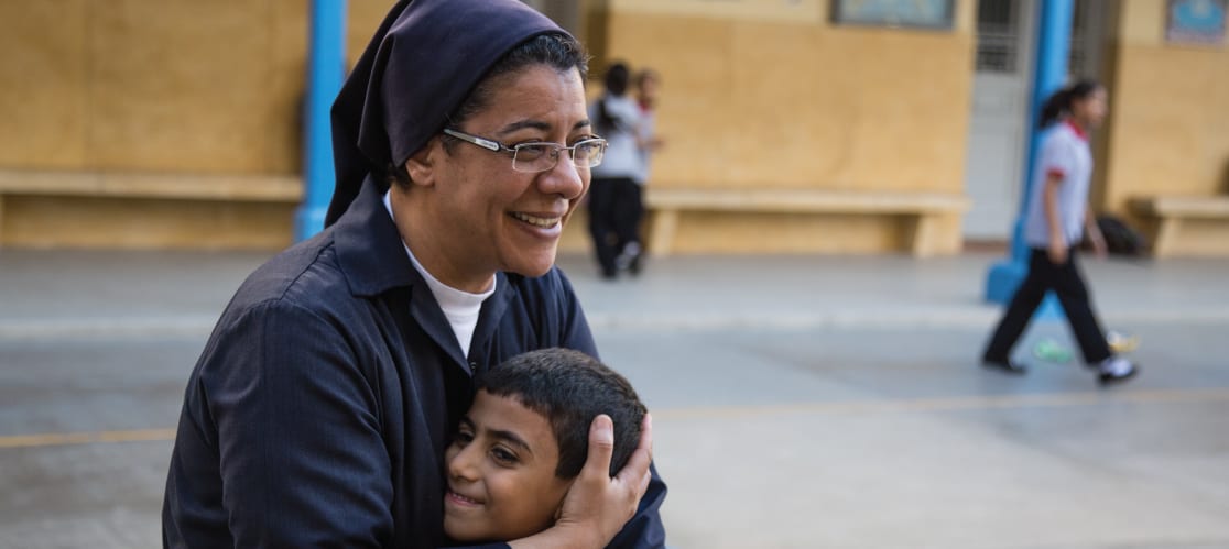 Nun hugging child in public setting, both smiling.