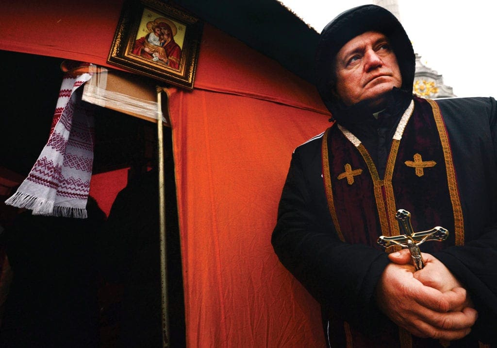 Hooded priest stands outside of a tent holding a cross.