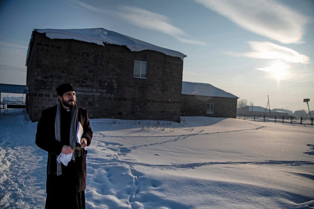 Priest standing in snow on path outside of church.