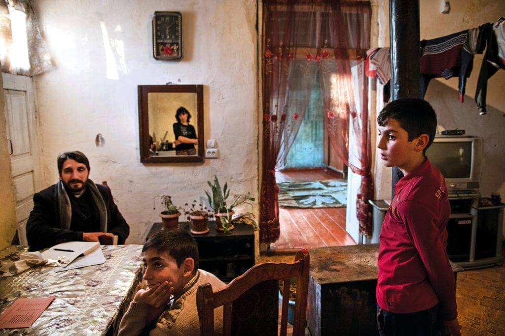 Priest sits in parishioners' home, chatting.