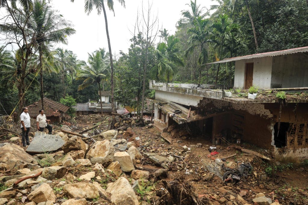 Distant figures of two priests standing amid flood debris outside a house in Kerala, India.