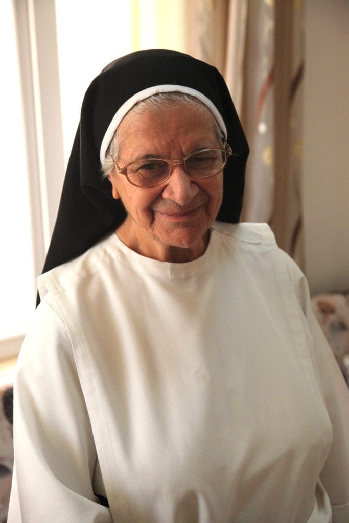 an elderly nun stands smiling in a well-lighted room.