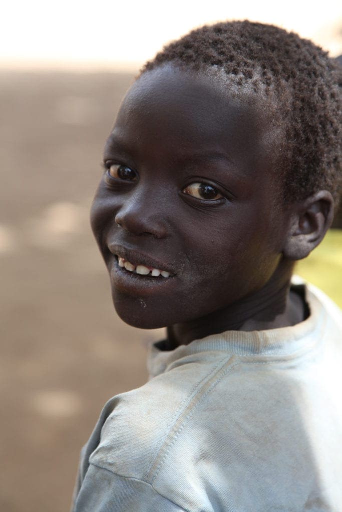 A young child smiles in the shade on a sunny day.