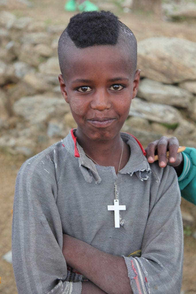 a child smiles, arms folded with a cross visible around his neck.