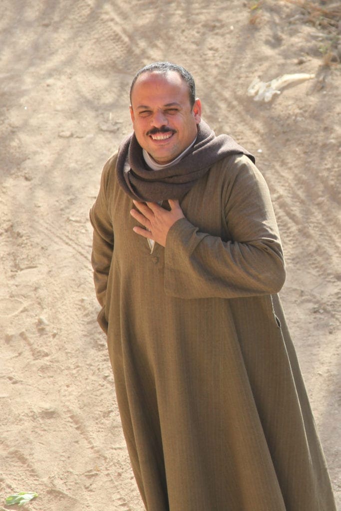 a man in robes smiles while standing on a dirt road.