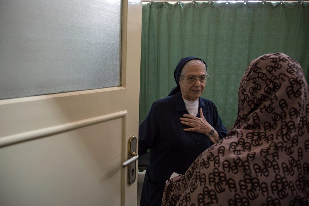 Sister Simone stands at a doorway and greets a woman in a headscarf.