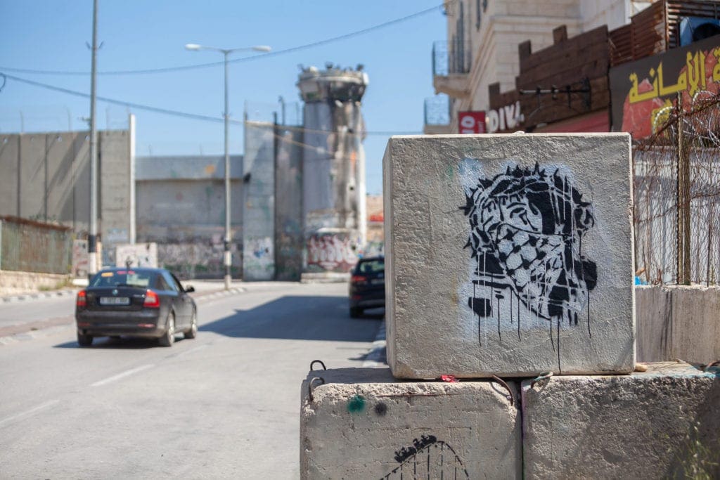 graffiti of Jesus wearing a mask is visible on a concrete block before an empty street.
