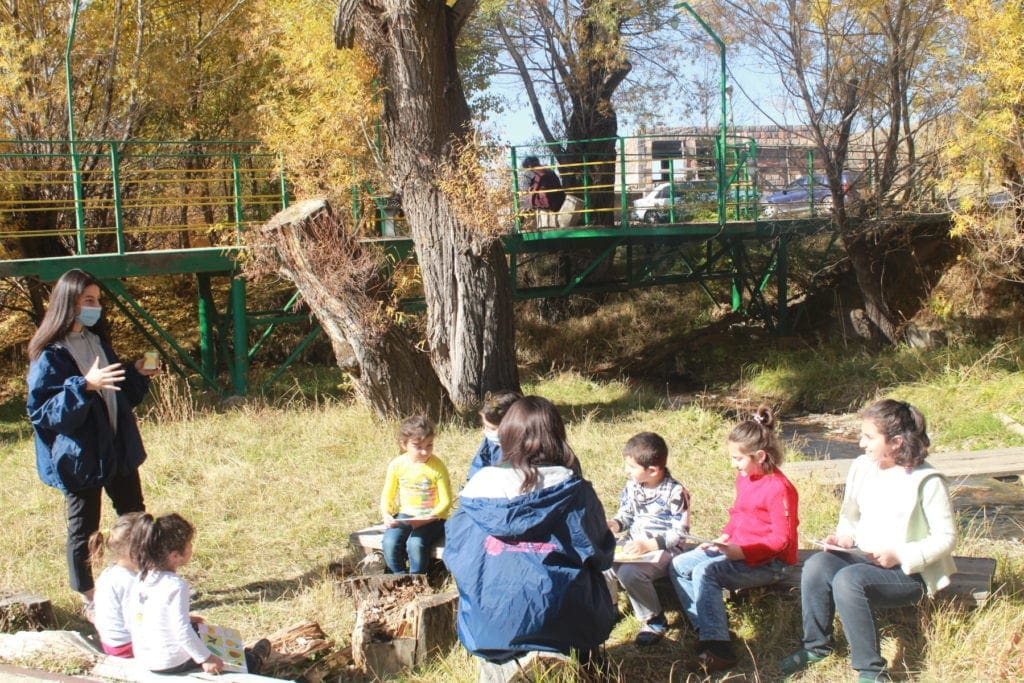 young women in masks speak to people, mostly children, seated outside.