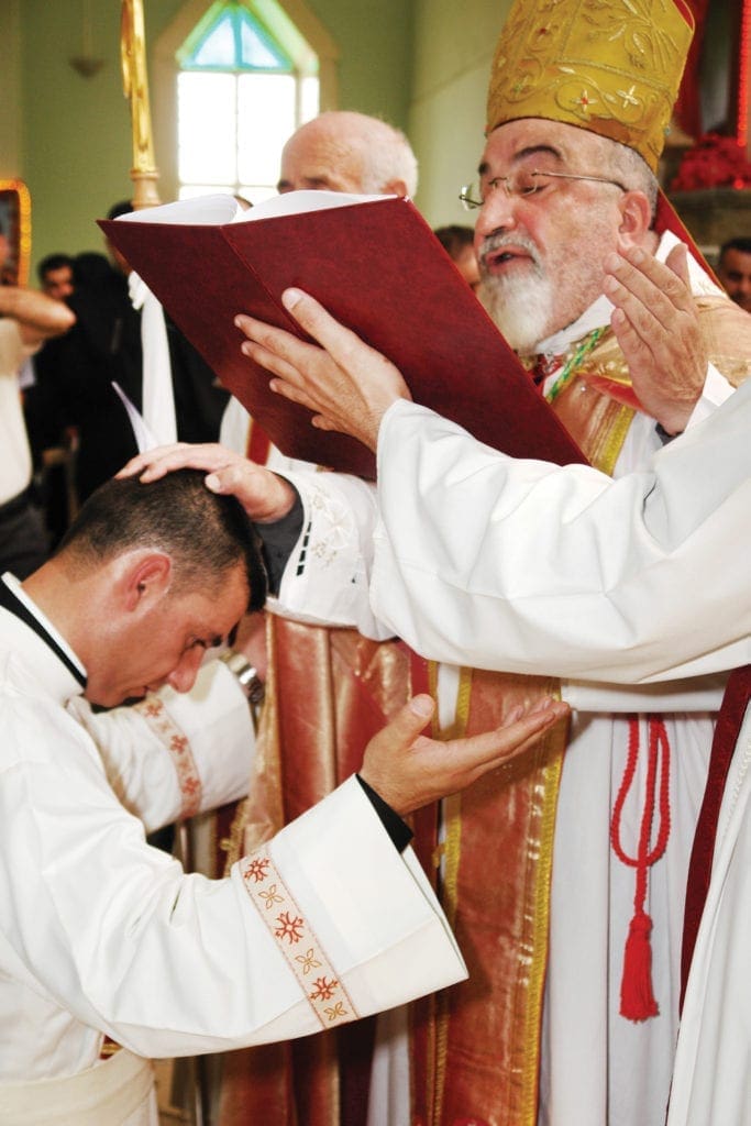an archbishop prays over the bowed head of a priest.