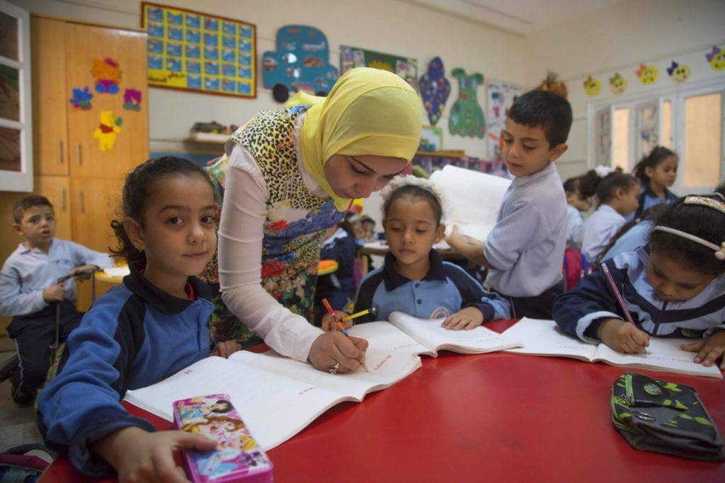 a teacher assists students with an assignment, writing in an open notebook at a table.