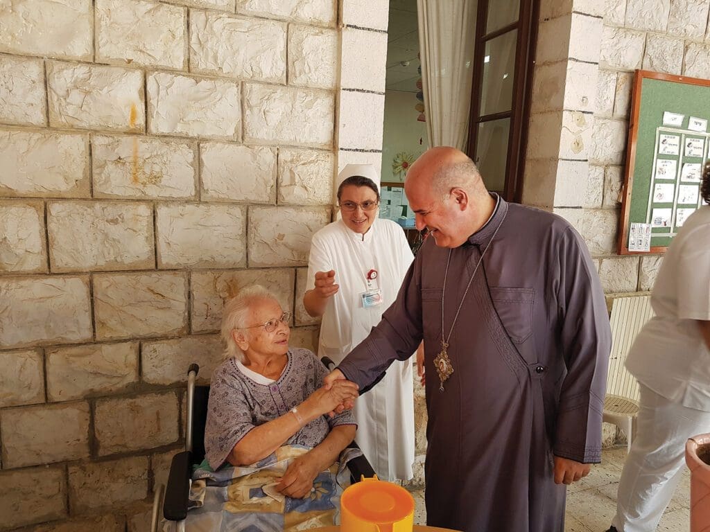 Archbishop Georges Bacouni greets an elderly woman in a wheelchair.