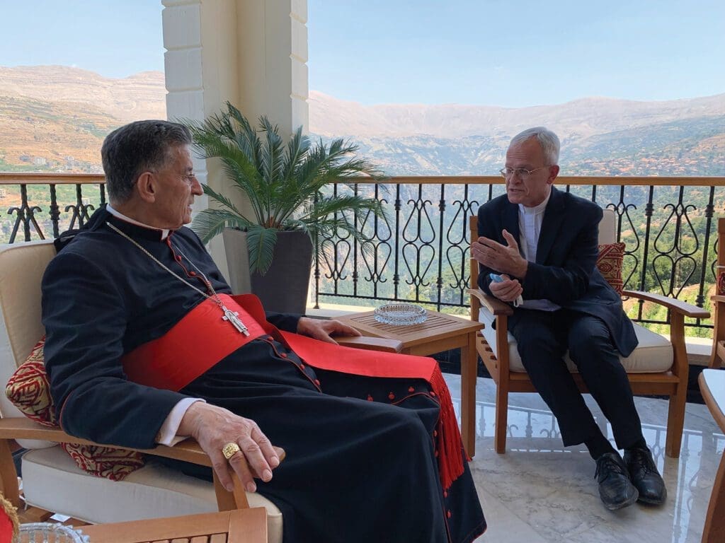 two men, seated, in clerical attire, talk outside on a balcony overlooking a hilly landscape.