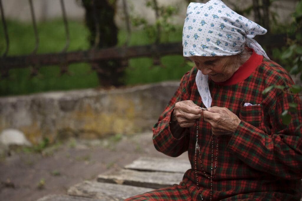An elderly woman in a red dress and white headscarf holds rosary beads.