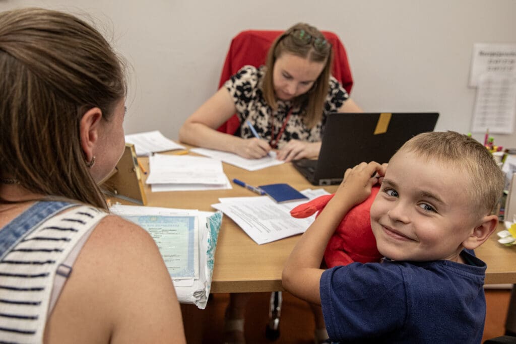 A child looking at the camera smiles while a woman sitting on a desk writes on a piece of paper.