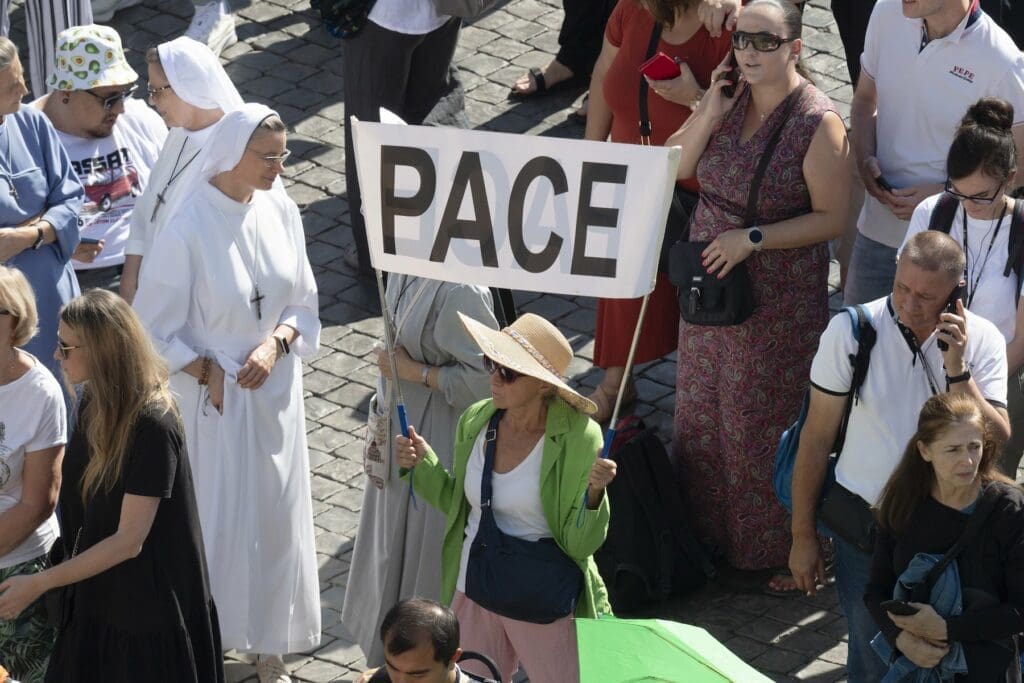 a woman holds up a white sign that reads "Pace". There are people standing near her, including a nun.