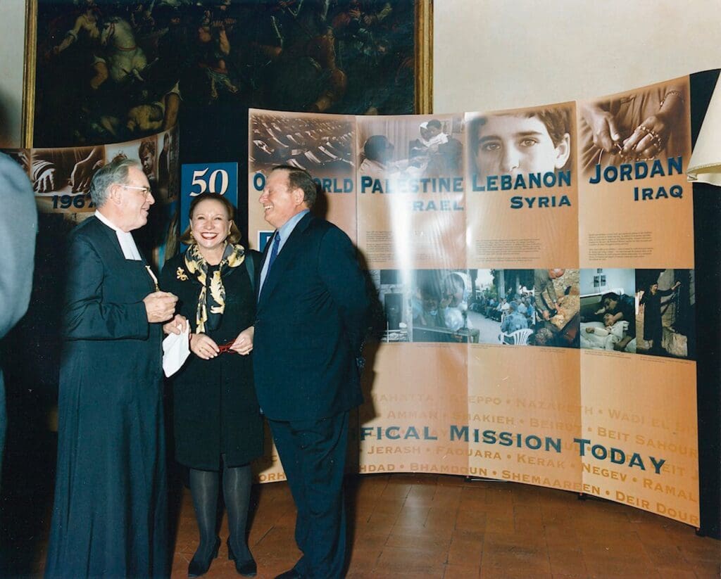 A man with a habit, a woman and another man smile while having a conversation in front of a panel with pictures from the Middle East.