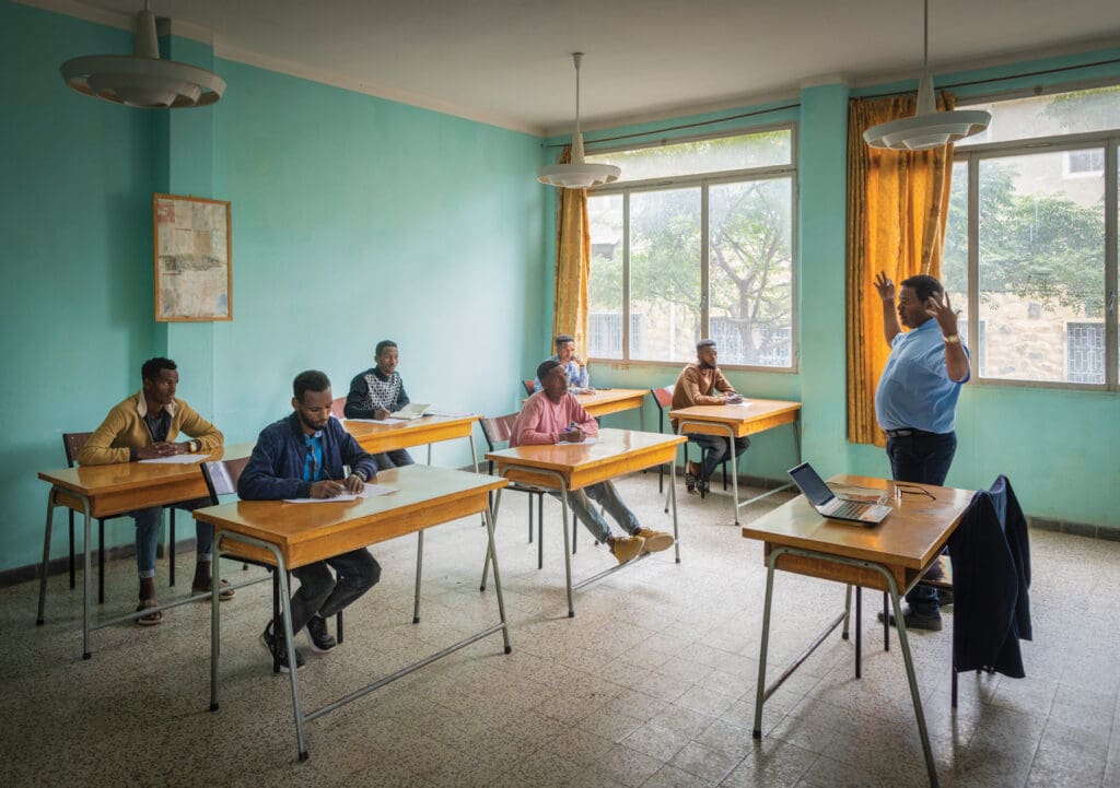A man, a teacher, explains something to six students sitting at desks.